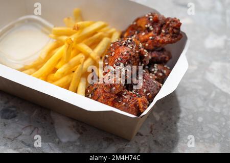 Amerikanische Küche. Essenslieferung, Essen zum Mitnehmen, Papierbehälter mit gebratenen Hähnchenflügeln mit Tomatensoße und pommes frites. Menü, Nahaufnahme Stockfoto