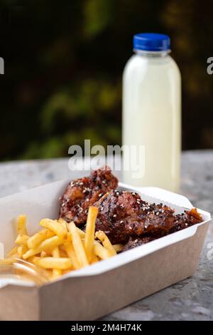 Amerikanische Küche. Essenslieferung, Essen zum Mitnehmen, Papierbehälter mit gebratenen Hähnchenflügeln mit Tomatensoße und pommes frites. Menü, Nahaufnahme Stockfoto