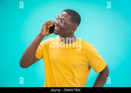 Attraktiver schwarzer Afrikaner, der am Handy spricht, glücklich lächelt, isoliert auf blauem Hintergrund. Stockfoto