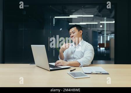 Ein junger Asiate sitzt im Büro am Tisch und arbeitet an einem Laptop. Er winkt ihm mit der Hand ins Gesicht. Er fühlt sich schlecht, fühlt sich heiß, braucht frische Luft, medizinische Hilfe. Stockfoto