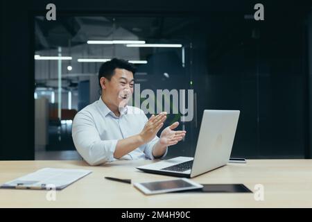 Geschäftstreffen. Ein junger, asiatischer Geschäftsmann veranstaltet ein Online-Geschäftstreffen, eine Konferenz. Sitzen am Tisch im Büro, schauen auf den Laptop, applaudieren, danken, lächeln. Stockfoto