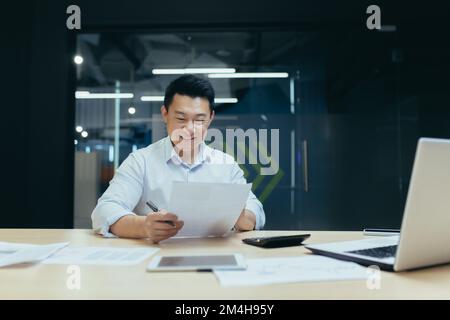 Glücklicher junger asiatischer Buchhalter, der an einem Schreibtisch im Büro sitzt, Dokumente in der Hand hält, einen Taschenrechner benutzt. Arbeitet mit Konten, Budget, Darlehen und Dokumenten. Vereinbarungen. Stockfoto