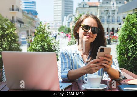 Junge Frau, die ihr Smartphone benutzt, während sie in einem Café im Freien in der Stadt sitzt. Stockfoto
