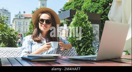 Junge Erwachsene Frau mit Sonnenbrille und Strohhut, die im Café im Freien sitzt und während der Geschäftsreise Kaffee trinkt. Stockfoto
