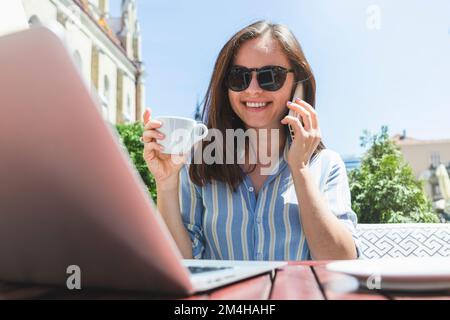 Junge lächelnde Frau, die im Sommer im Café im Freien sitzt und Kaffee trinkt, über ein Handy spricht und auf einen Laptop schaut. Stockfoto