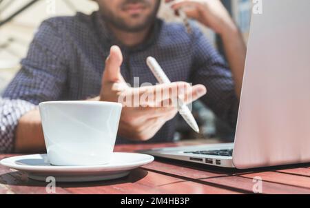 Weiße Tasse Kaffee auf dem Tisch und unscharfer Mann, der im Hintergrund an einem Laptop arbeitet. Stockfoto
