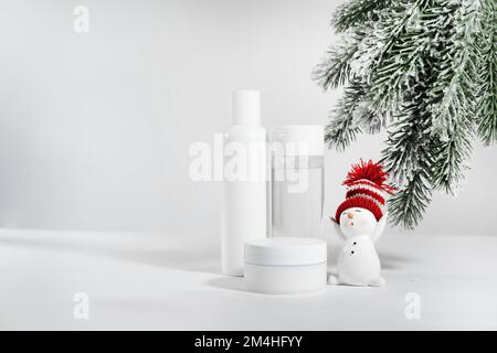 Weißes Gefäß mit Hautpflegekosmetika auf einer weißen Oberfläche mit einem Schneemann. Winterkosmetik. Leerer Container mit Tannenzweigen im Hintergrund. Weihnachtsgeschenk ide Stockfoto