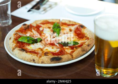 Heiße verführerische Pizza und frisches Glas Bier auf einem weißen Teller auf einem Holztisch. Seitenansicht einer köstlichen, dünnen, käsigen Pizza. Italienische Küche Stockfoto