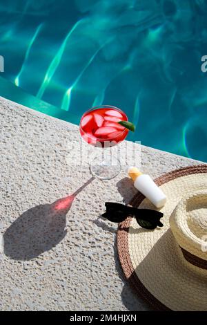 Erfrischender roter Cocktail am Pool mit Sonnencreme, Sonnenbrille und Strohhut Stockfoto