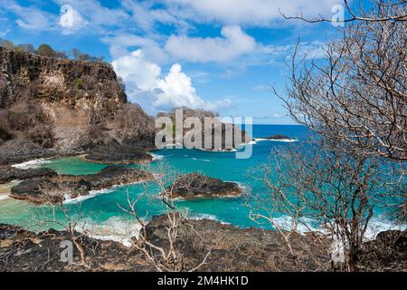 Blick auf eine der kleinen Buchten von Fernando de Noronha mit kristallklarem Wasser und kleinen felsigen Inseln mit trockener Vegetation im Vordergrund Stockfoto