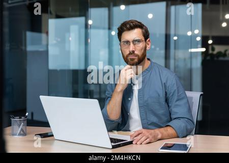 Ein seriöser, nachdenklicher Geschäftsmann im Hemd, der über eine Entscheidung nachdenkt, sitzt am Tisch im modernen Büro und ein Mann mit Bart benutzt bei der Arbeit ein Notebook. Stockfoto