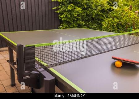 Blick auf Tischtennisschläger und orangefarbene Plastikkugel auf einem grünen Tennistisch mit Netz im Innenhof der Villa. Konzept des aktiven Lebensstils. Schweden. Stockfoto