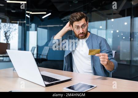 Frustrierter und betrogener Geschäftsmann im Büro, der im Online-Shop einkaufen und Geld per Bankkreditkarte überweisen wollte, genervter Mann, der im Büro mit Laptop und Telefon arbeitet. Stockfoto