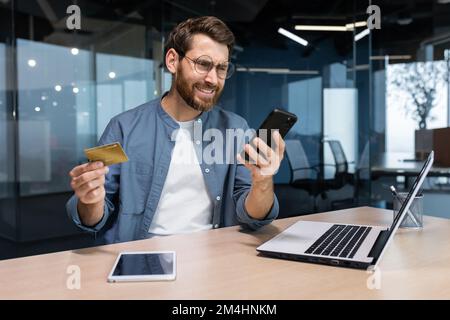 Frustrierter und betrogener Geschäftsmann im Büro, der im Online-Shop einkaufen und Geld per Bankkreditkarte überweisen wollte, genervter Mann, der im Büro mit Laptop und Telefon arbeitet. Stockfoto