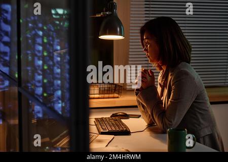 Junge asiatische Geschäftsfrau, die an ihrem Arbeitsplatz vor dem Computer sitzt und Überstunden im Büro macht Stockfoto