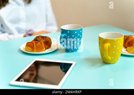 Croissants und Kaffeetassen für zwei Personen auf einem Tisch mit einem Tablet. Verschwommenes Bild eines Mädchens, das auf dem Stuhl sitzt. Blaue und gelbe Kaffeetassen mit weißer Polka Stockfoto