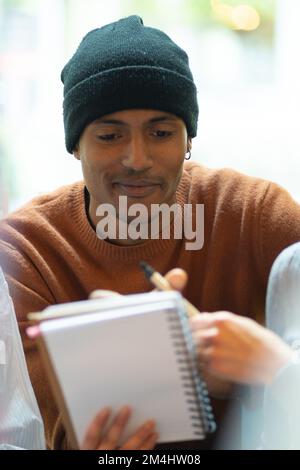 Kollegen, die in einem Teamarbeitsbereich zusammenarbeiten. Dunkelhäutiger Typ mit schwarzer Mütze und Pullover. Ein Mädchen mit einem Stift in der Hand zeigt etwas auf dem Stockfoto