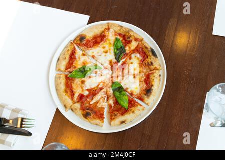 Heiße, verführerische Pizza auf einem weißen Teller auf einem Holztisch. Draufsicht auf eine köstliche, käsige Pizza mit Gabeln, Messer und Servietten auf dem Tisch. Dünn Stockfoto