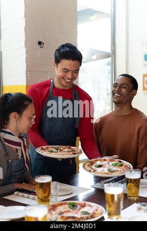 Der Kellner serviert einer Gruppe von Freunden köstliche Pizza in einem zwanglosen Café. Der Kellner hielt zwei Pizzen in der Hand und legte sie auf den Tisch. Drin Stockfoto