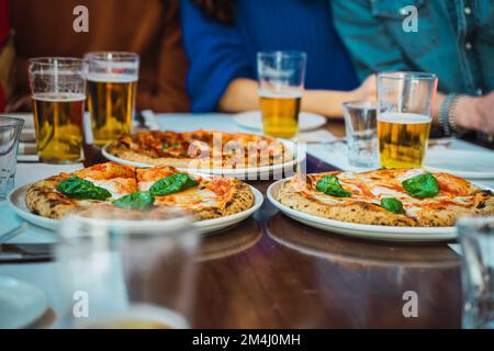 Pizza und Bier in Gläsern auf einem Holztisch. Freunde feiern Pizza nach der Arbeit. Nahaufnahme von drei Pizzas auf weißen Tellern. Mittagessen bei der Arbeit. Shar Stockfoto