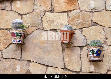 Pflanzmaschinen mit Kaktus, die an einer Steinwand hängen. Rustikal, große Steine, Lehm, Schlamm, Dekoration, Metallhalterungen, Vorderansicht, Vorderansicht Stockfoto