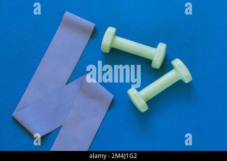 Draufsicht auf Kurzhanteln und Fitnessband auf blauer Sportmatte. Flach liegend, Kopierbereich. Heimtrainingkonzept Stockfoto