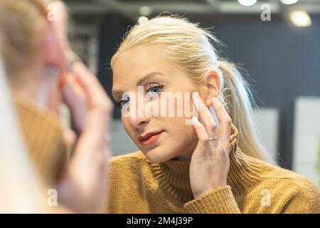 Blondes Mädchen vor dem Spiegel, das Akne auf ihrer Gesichtshaut prüft. Versorgungskonzept für Haut und dermatologische Jugendstörungen wie Pickel Stockfoto