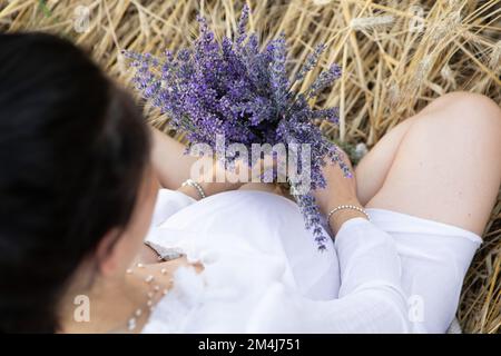 Nahaufnahme des Bauches einer schwangeren Frau in einem Weizenfeld mit Lavendel in der Nähe ihres Bauches. Unbekanntes, anonymes Gesicht. Stockfoto