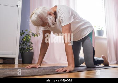 Seniorin, die Yoga im Zimmer macht, mit Blick auf die Katze Stockfoto