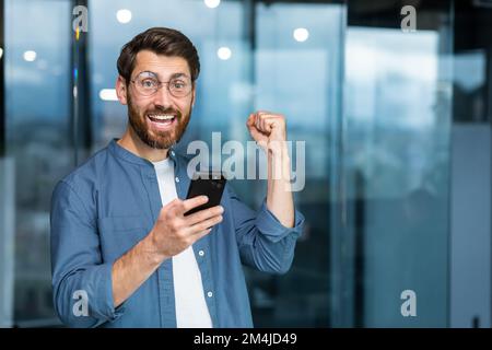 Porträt eines modernen Geschäftsmannes mitten im Büro, der Mann schaut mit einem Smartphone in die Kamera und feiert den Sieg, der Mann hält das Telefon in den Händen und hebt seine Hand in einer Geste des Triumphes. Stockfoto