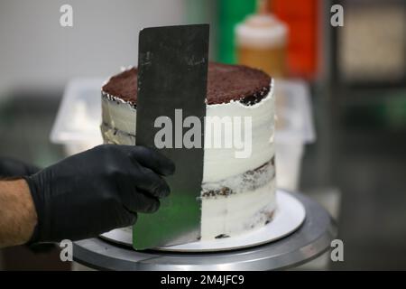 Der Konditor verwendet einen Spatelkratzer, um eine große, zilindrikale, matte Torte mit Buttercreme zu glätten – Zubereitung von Hochzeitstorten Stockfoto