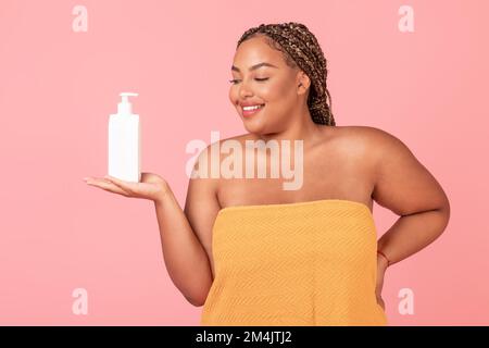 Wunderschöne schwarze, übergroße Frau, die eine Flasche mit Körperlotion hält und ein neues Hautpflegeprodukt empfiehlt Stockfoto