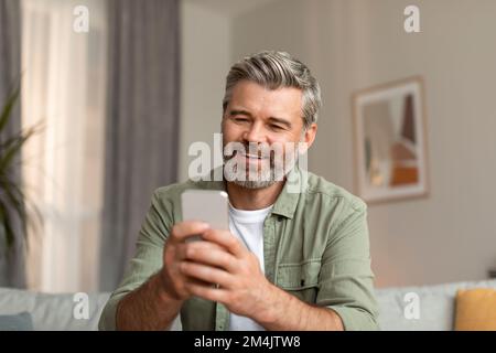 Lächelnder älterer, weißer Blogger, männlicher Blogger, der auf dem Smartphone tippt, verwendet eine neue App für Chat und Video Stockfoto