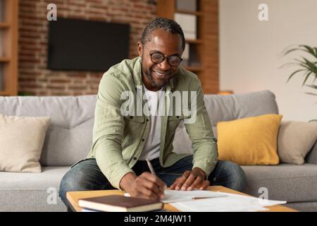 Ein fröhlicher schwarzer Geschäftsmann mit Brille macht Notizen, lernt und arbeitet im Wohnzimmer, im Kopierraum Stockfoto