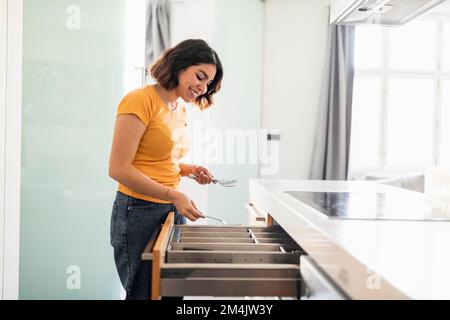 Lächelnde Frau Aus Dem Nahen Osten, Die Besteck In Der Küchenschublade Aufräumt, Seitenansicht Stockfoto