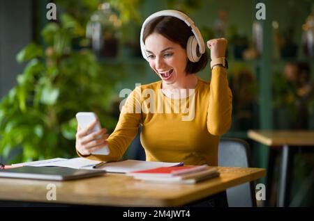 Emotionale junge IT-Frau, die Kopfhörer benutzt, hört Musik am Telefon Stockfoto
