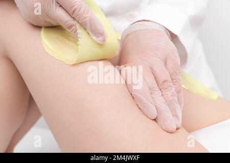 Nahaufnahme von Kosmetikhänden, die Haarentfernung an weiblichen Beinen mit grünem heißem Wachs im Schönheitssalon vornehmen. Der Wachsvorgang der Haarentfernung aus der Wurzel Stockfoto
