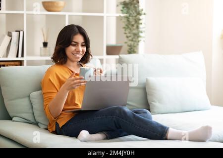 Lächelnde Junge Arabische Frau, Die Sich Mit Laptop Und Kaffee Auf Dem Sofa Ausruht Stockfoto