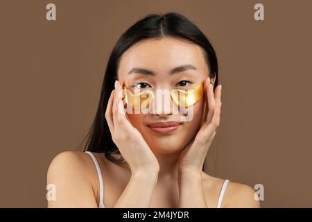 Glückliche koreanische Frau, die Augenklappen anbringt, sich auf braunem Hintergrund operiert, Studioaufnahme Stockfoto