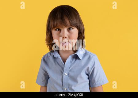 Kindliche Verwöhnung. Süßer kleiner Junge, der rummacht, seine Zunge rausstreckt und seine Augen schielt, orangefarbener Hintergrund Stockfoto