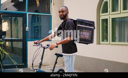 Ein lächelnder afroamerikanischer Mann, der draußen auf Kunden wartet, fährt mit dem Fahrrad mit Thermal-Rucksack, um Essen zum Mitnehmen zu liefern. Der Kurier steht neben dem Eingang zum Bürogebäude und sieht in die Kamera. Stockfoto
