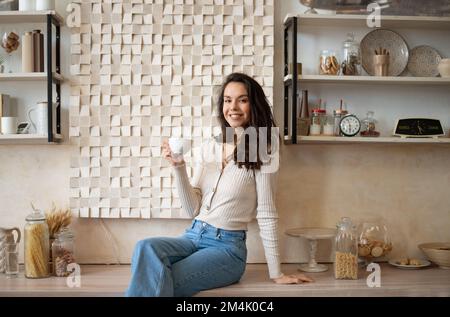 Sorgenfreie, glückliche Dame, die sich in der gemütlichen Küche entspannen kann, an der Theke sitzt und Kaffee trinkt, vor der Kamera lächelt, Kopierbereich Stockfoto