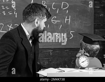 Ein junger männlicher Lehrer führt seinen Schüler zum Lernen, während der Sohn mit Kritzeleien auf der Tafel sitzt und im Klassenzimmer sitzt Stockfoto