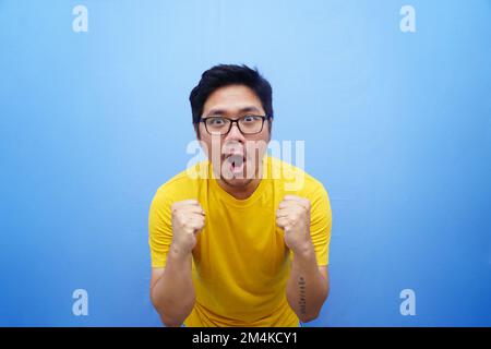 Ein junger asiatischer Mann mit einem glücklichen, erfolgreichen Gesichtsausdruck, der ein gelbes T-Shirt trägt, isoliert auf blauem Hintergrund Stockfoto
