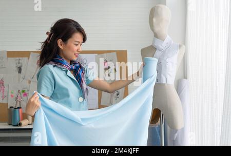 Modedesignerzimmer. Junger asiatischer Schneiderin in gestreiftem Schal, der die Qualität des blauen feinen Stoffes von der Rolle prüft. Stockfoto