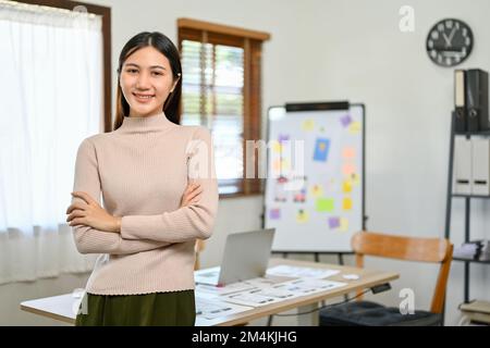 Eine attraktive und selbstbewusste Chefin der Millennials in Asien steht mit gekreuzten Armen, lächelt und schaut in die Kamera vor dem Konferenzraum. Stockfoto
