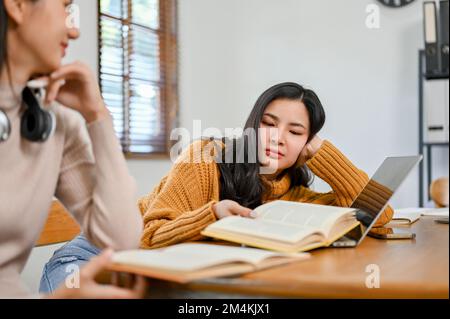 Gelangweilte, müde und schläfrige junge asiatische Studentin, die sich auf die Prüfung vorbereitet und ein Buch mit ihrer Freundin zu Hause liest. Stockfoto