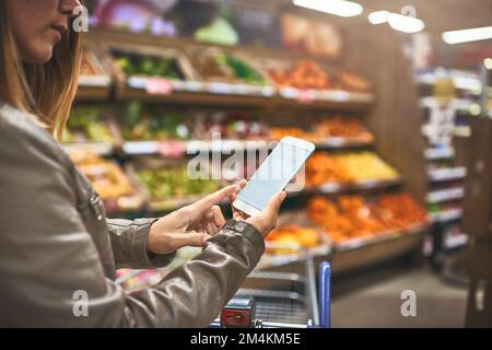 Sie behält alles im Auge, was sie mit Mobiltechnologie kauft. Eine Frau, die ein Handy in einem Supermarkt benutzt. Stockfoto