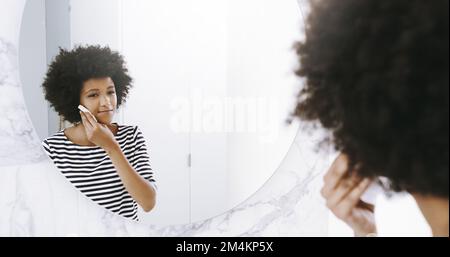 Ihr Tag beginnt mit dem Leuchten. Eine selbstbewusste junge Frau wischte ihr Gesicht mit einem Reinigungsmittel, während sie zu Hause in einen Spiegel im Badezimmer schaute Stockfoto