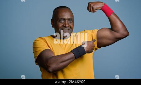 Ein lächelnder, erwachsener Sportler, der im Studio auf grauem Hintergrund steht und große männliche Muskeln zeigt, demonstriert die Pumps-Fitness eines männlichen Trainers Stockfoto